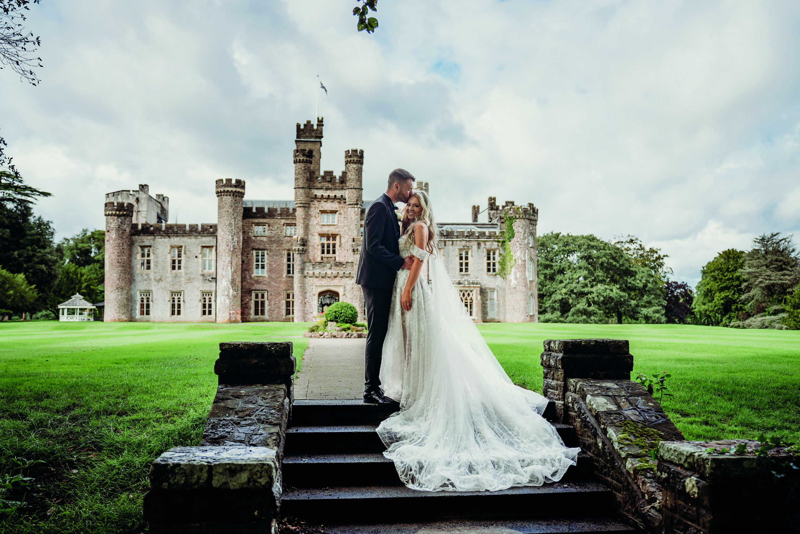 A bride and groom stand on stone steps in front of a large castle, surrounded by green lawns and trees, with a cloudy sky above.