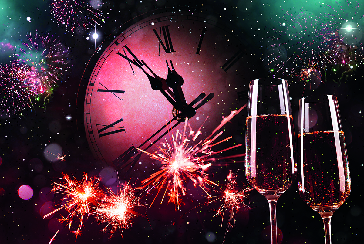 Fireworks illuminate the night sky with a large clock showing midnight in the background, alongside two filled champagne flutes celebrating the new year.