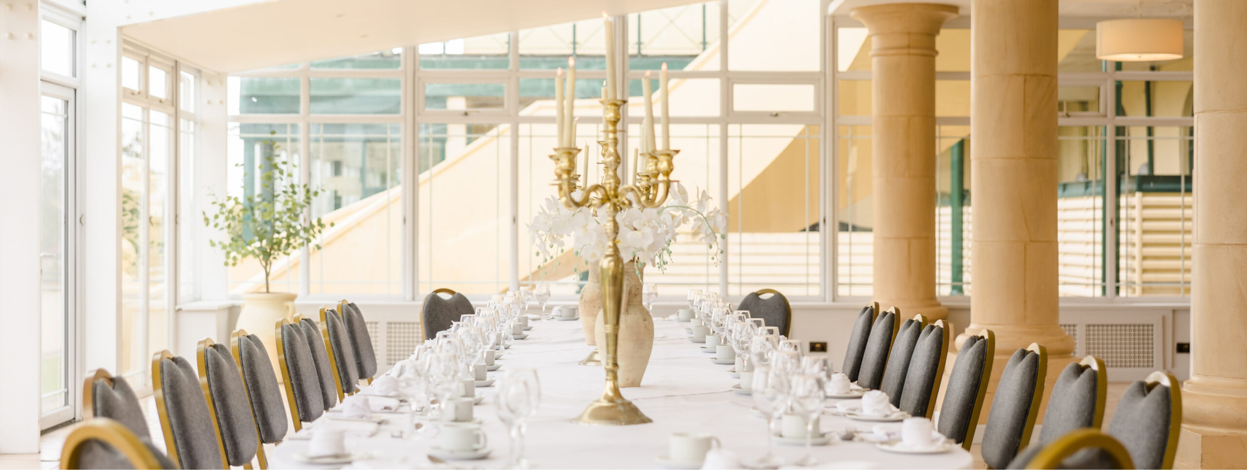 Elegant dining room with a long table set for an event, featuring a candelabra centerpiece, white tablecloth, china, and chairs in a well-lit space with large windows and columns.