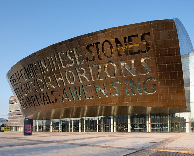 Modern building with a large bronze facade, featuring cut-out words in English and Welsh. Clear blue sky in the background.