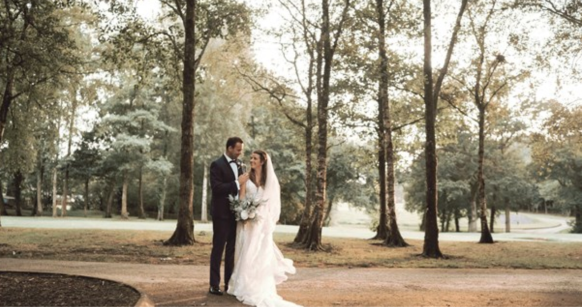 A bride and groom stand closely together on a tree-lined path, with the bride in a white dress and veil holding a bouquet, and the groom in a dark suit.