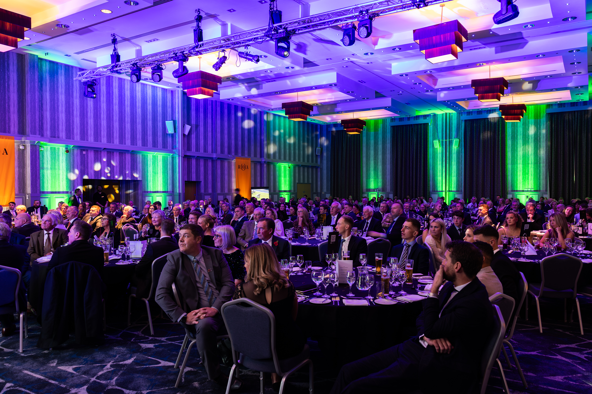 A large, formal banquet event with guests seated at round tables in a dimly lit room with green and purple lighting.