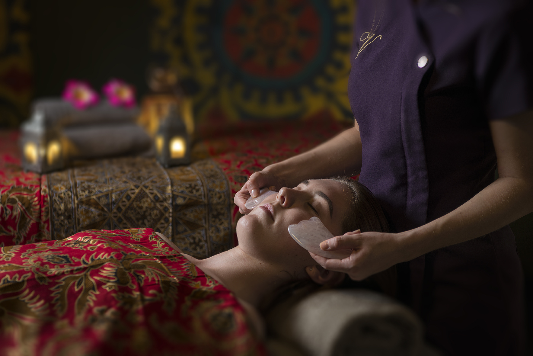 A person receiving a relaxing facial massage with stones, lying on a patterned bed, surrounded by soft lighting and decor.