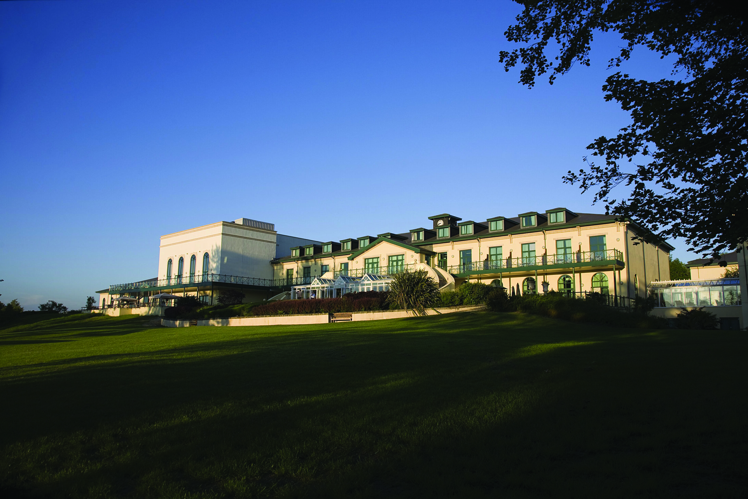 A large building with green rooftops and multiple windows sits on a grassy hillside under a clear blue sky. Trees are partially visible on the right.