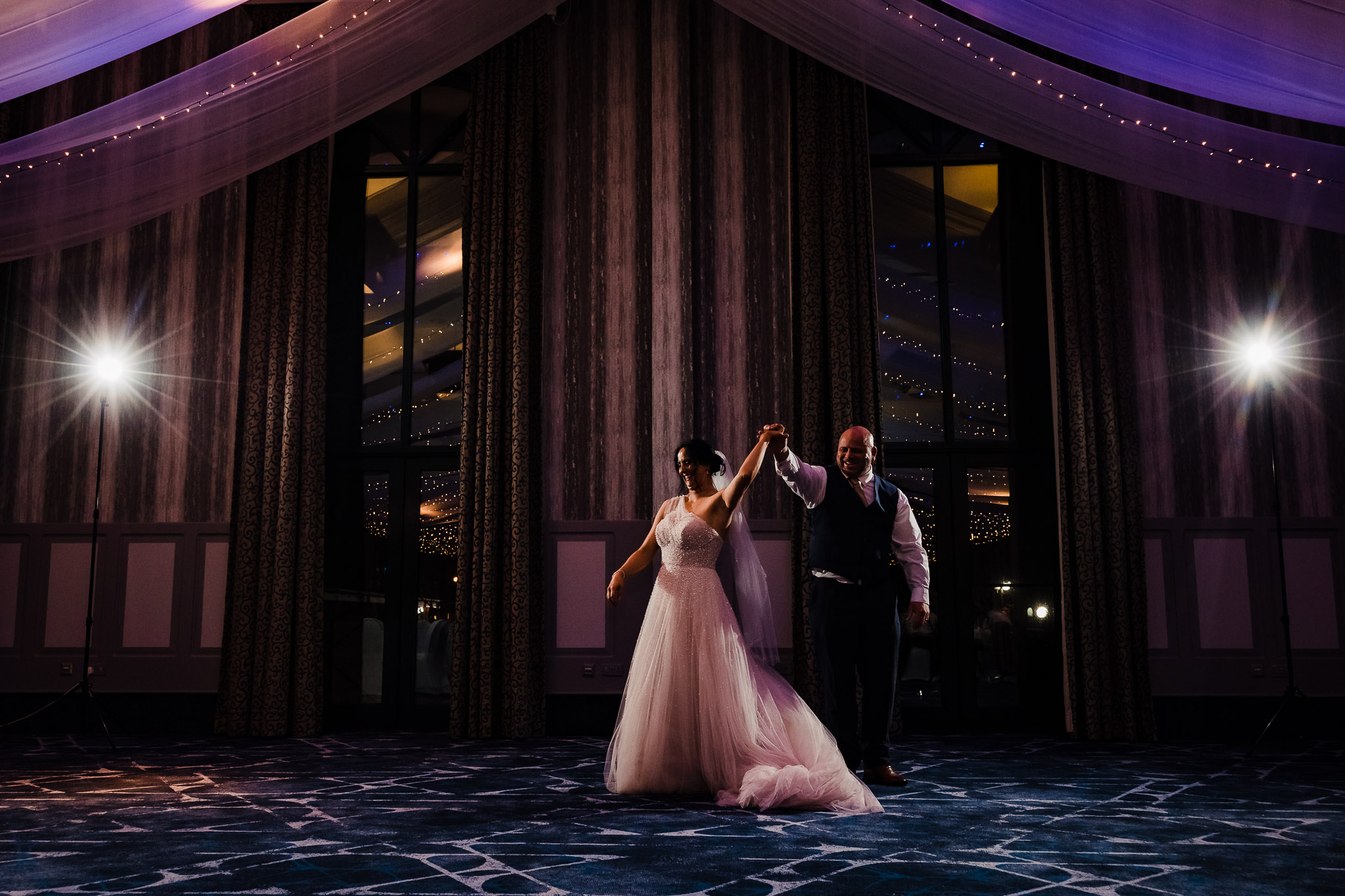 A bride and groom perform their first dance in a dimly lit ballroom with draped fabric and bright spotlights.