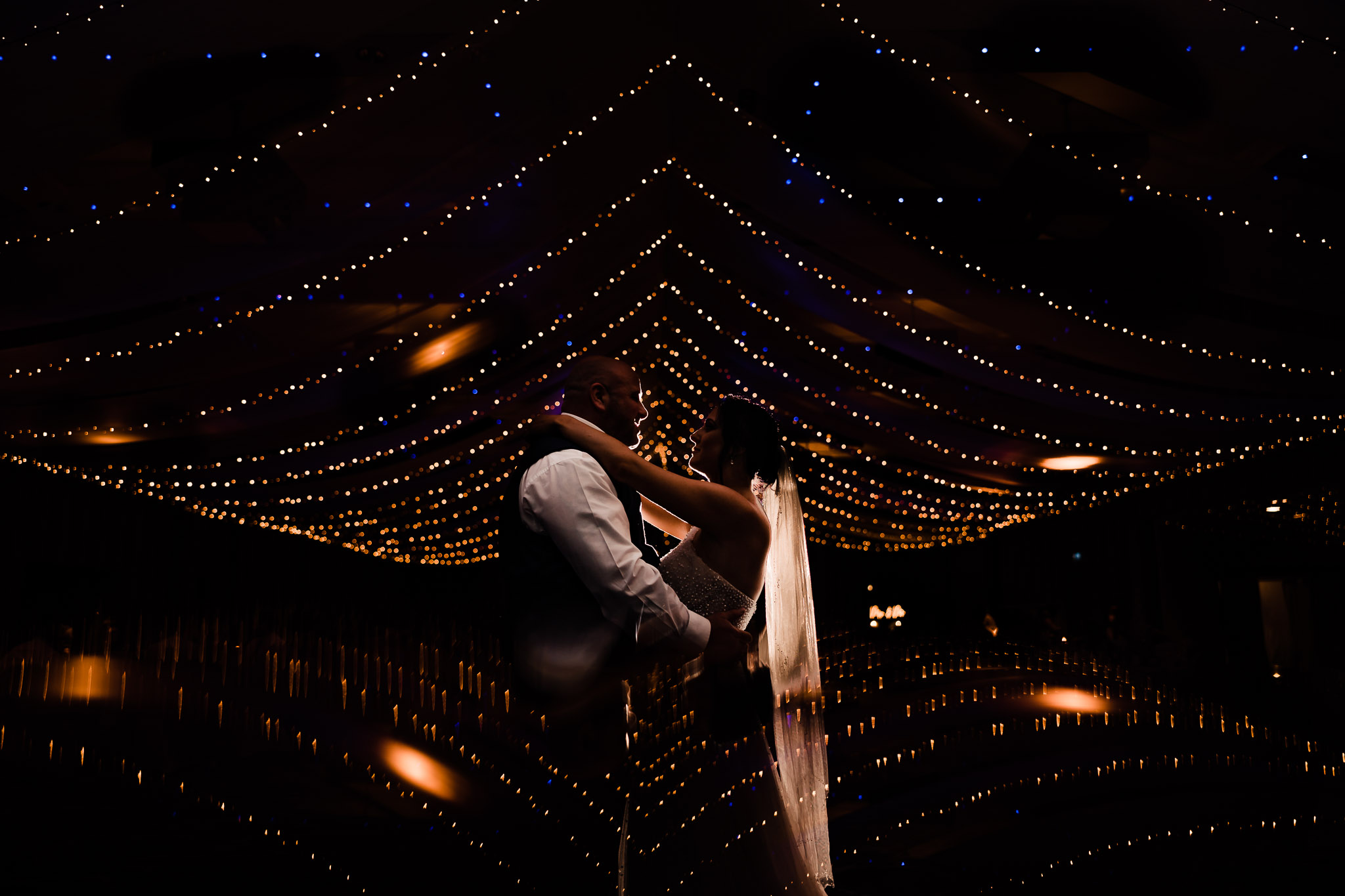 A couple dances beneath strings of twinkling lights in a dimly lit room.