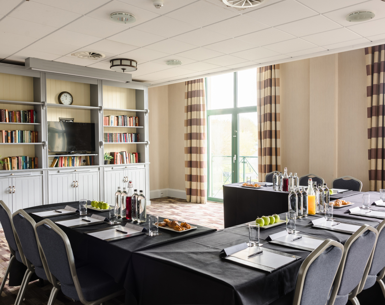 A conference room with U-shaped table setup, black tablecloths, bottled water, juices, pastries, and apples. Shelves with books and a TV are in the background. Natural light streams in.