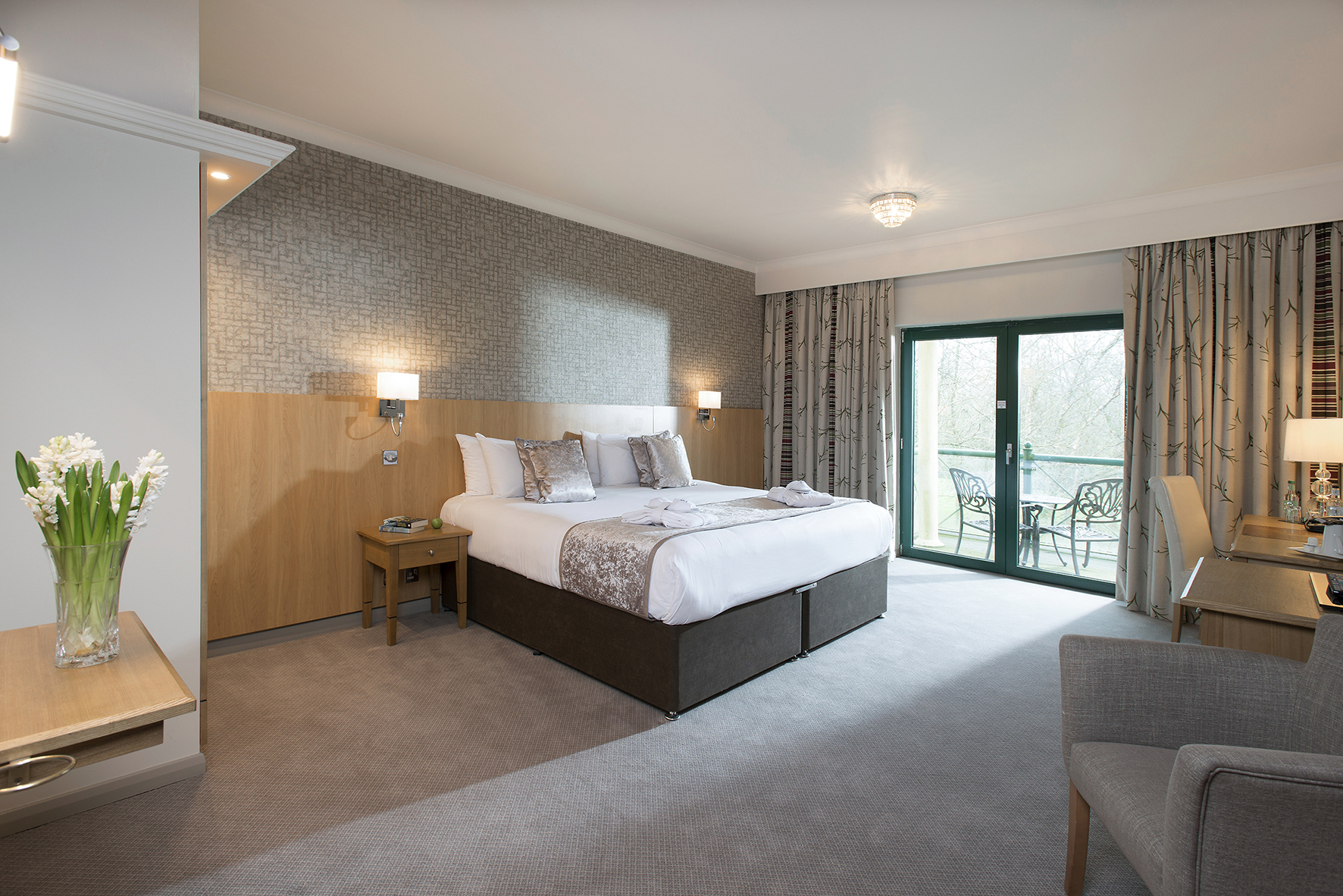 A hotel room with a large bed, a small table, a vase of flowers, a gray armchair, and a balcony with glass doors.