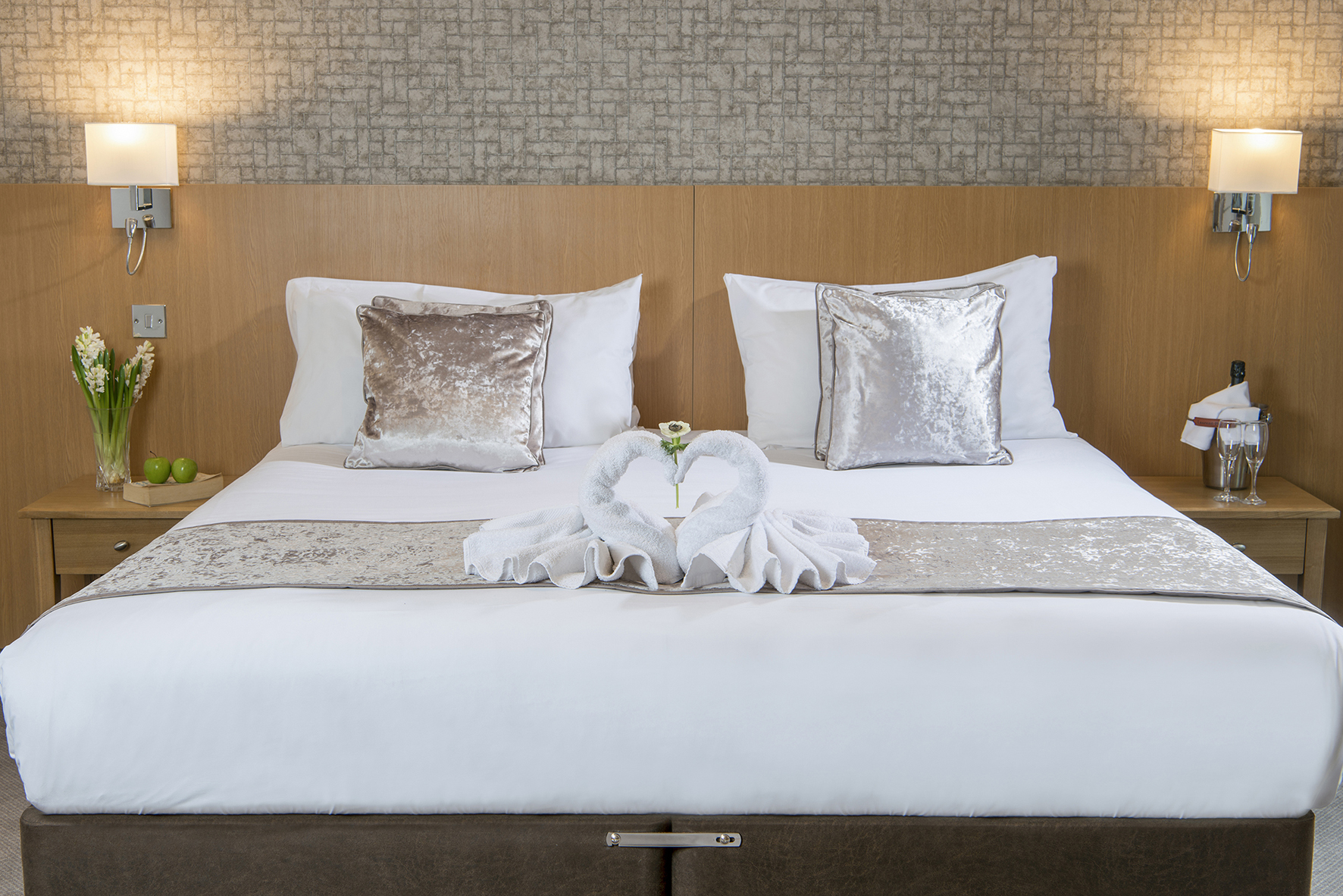 Hotel room with a neatly made double bed, silver pillows, swan-shaped towels, a small flower, and bedside tables with lights and decor items.