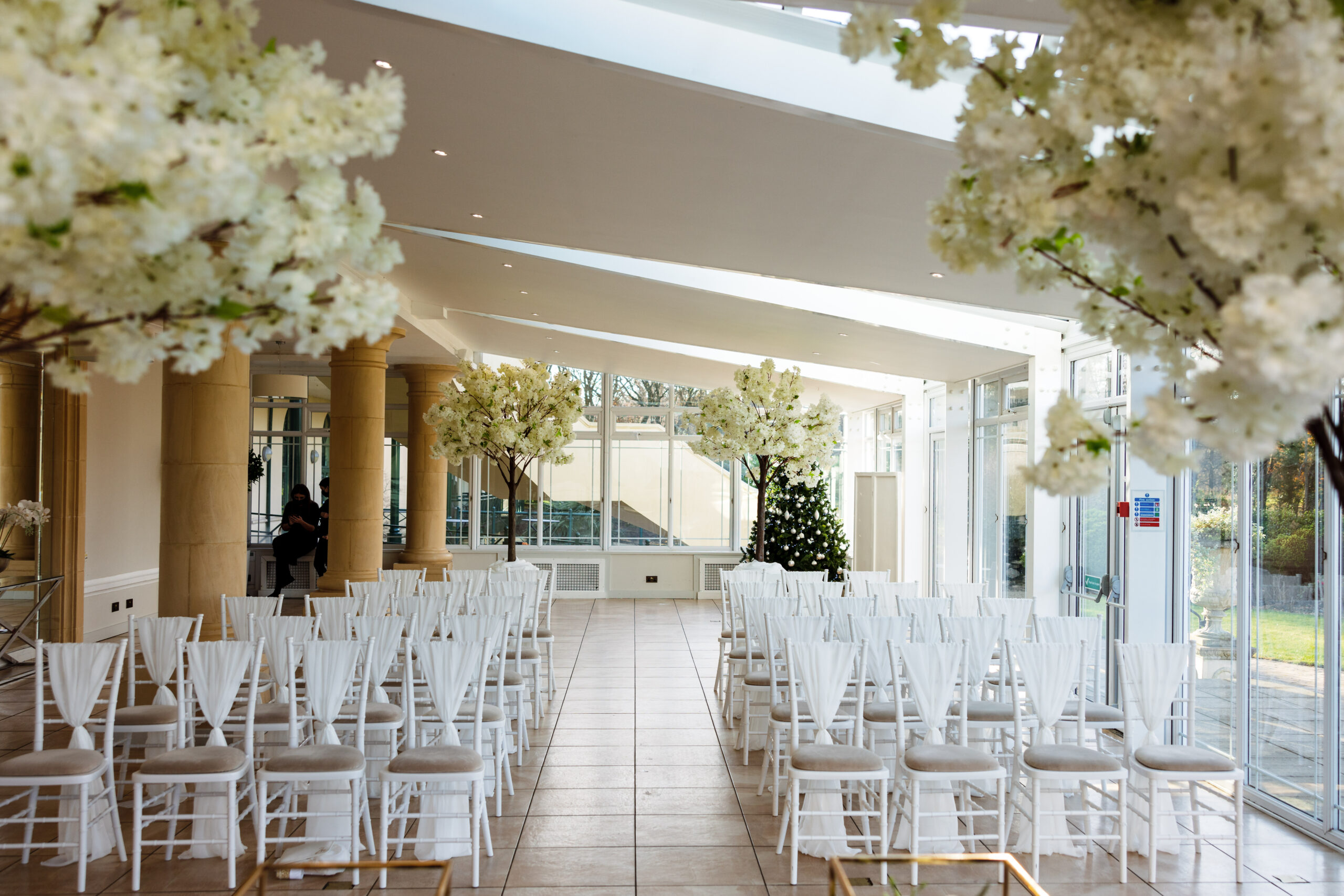 A bright wedding venue with rows of white chairs facing an altar, surrounded by white floral decorations and large windows.