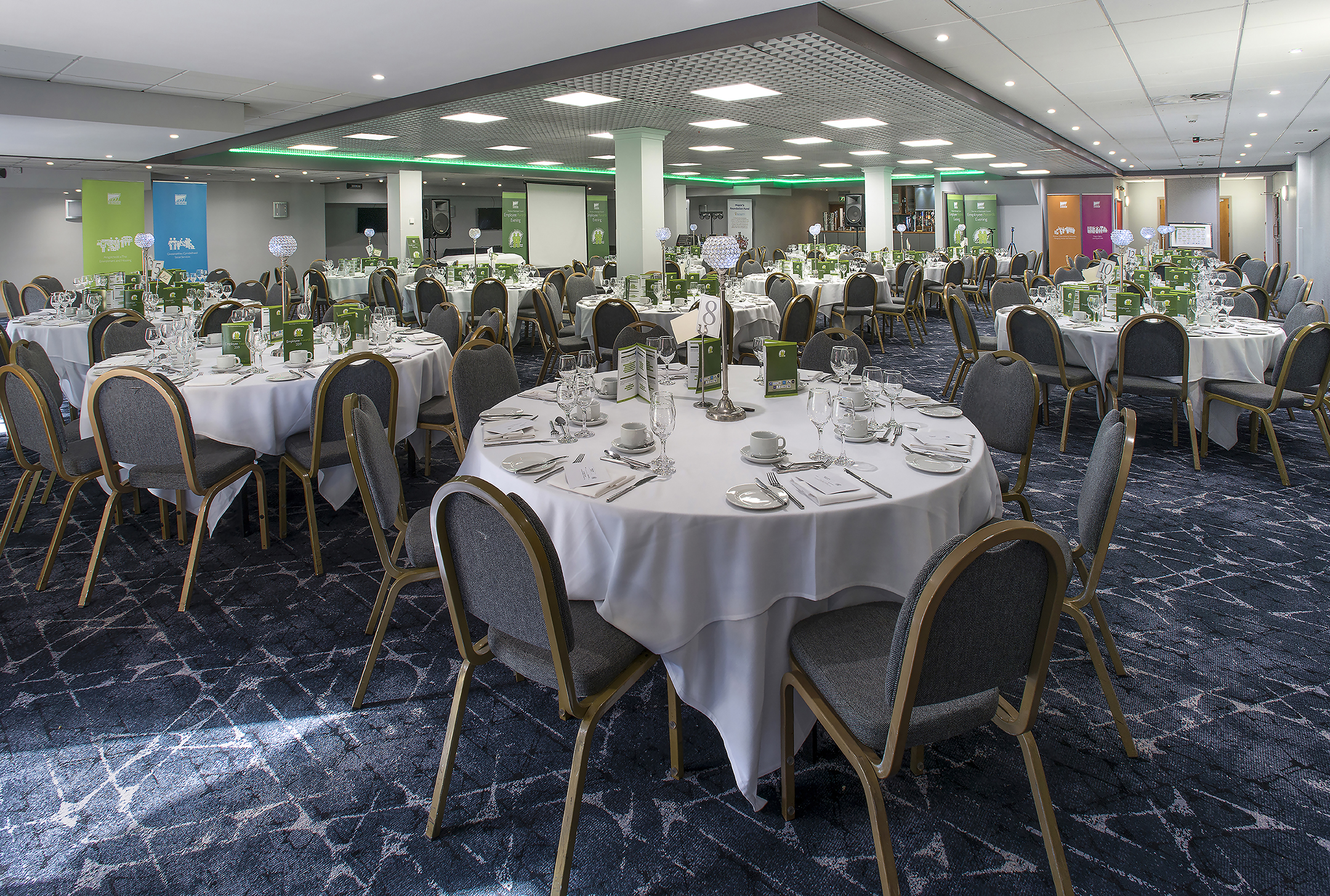 A large banquet hall with round tables set for dining, featuring white tablecloths, chairs, and decorative green accents.