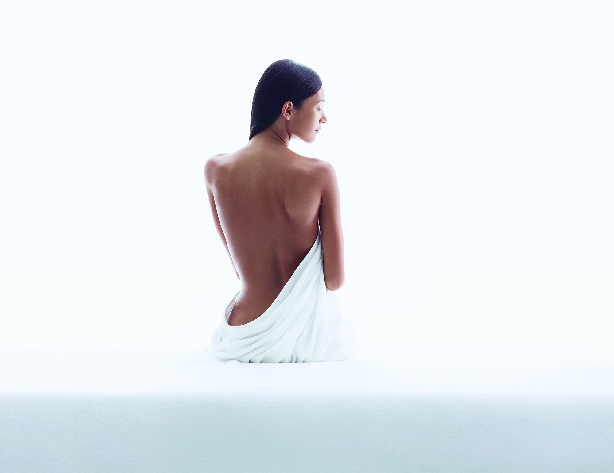 A woman with smooth skin sits with her back facing the camera, draped in a white cloth against a bright background.