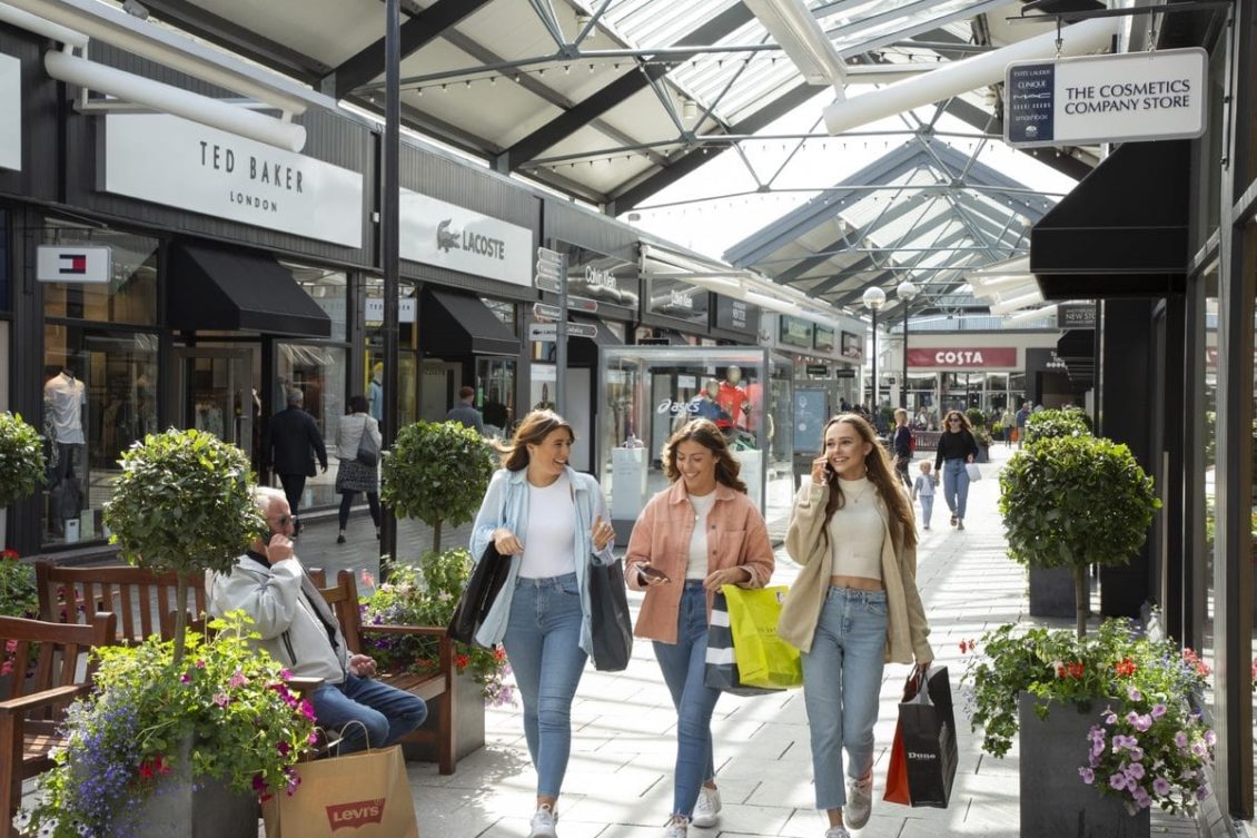 Three people walk with shopping bags in an outdoor shopping mall, with stores and a seated person in the background.