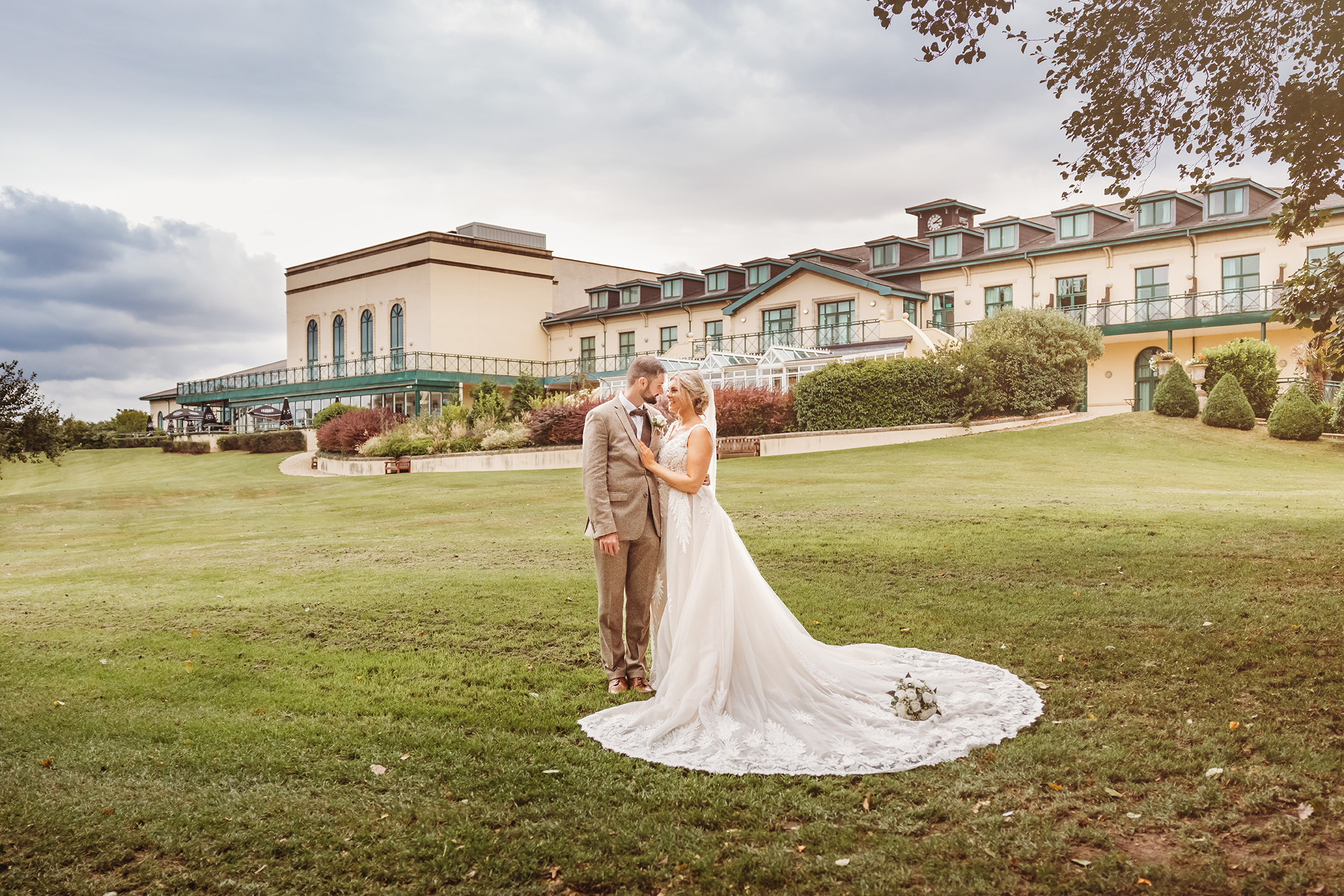 A bride and groom stand embracing on a green lawn, with a large building in the background, under a partly cloudy sky.