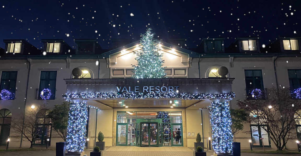 Entrance of Vale Resort decorated with festive lights and a tree. Snowflakes are falling in the evening sky.