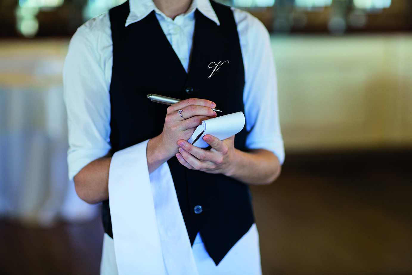 A server in a black vest and white shirt holds a notepad and pen, ready to take an order.