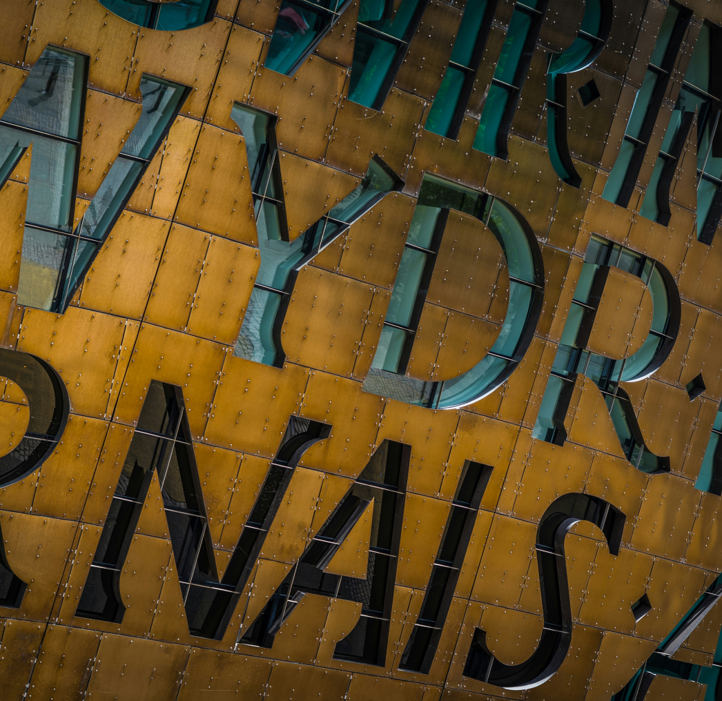 Close-up of a building facade with large, metallic lettering in two languages on a golden background. The letters appear to be part of a larger inscription.