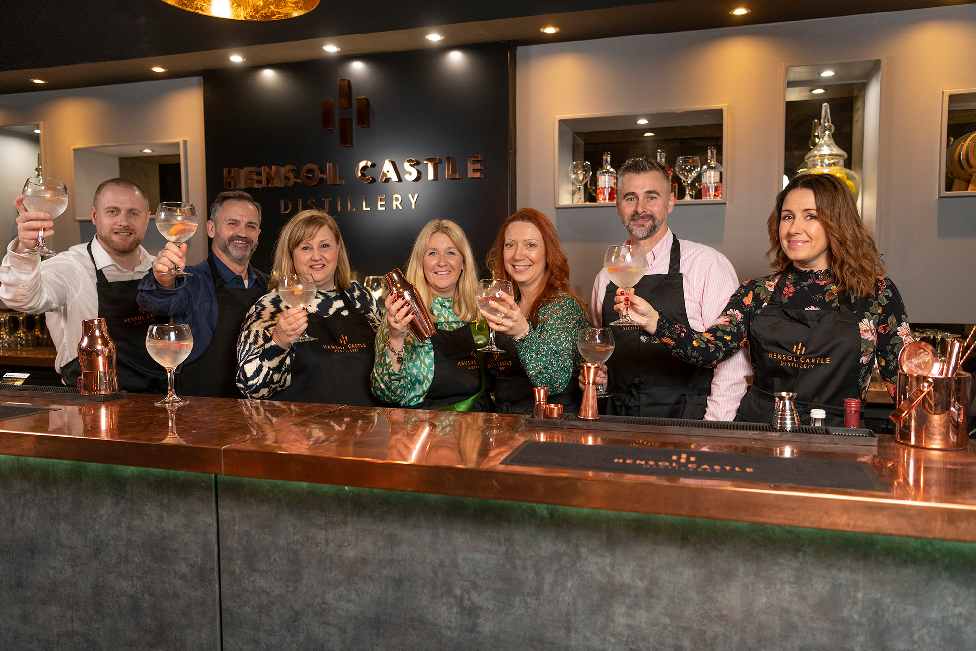 A group of people standing behind a bar at Henley Castle Distillery, holding glasses and smiling at the camera.