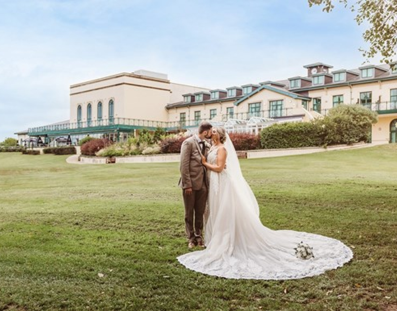 A bride and groom stand together on a lawn in front of a large building, the bride's long train spread on the grass.