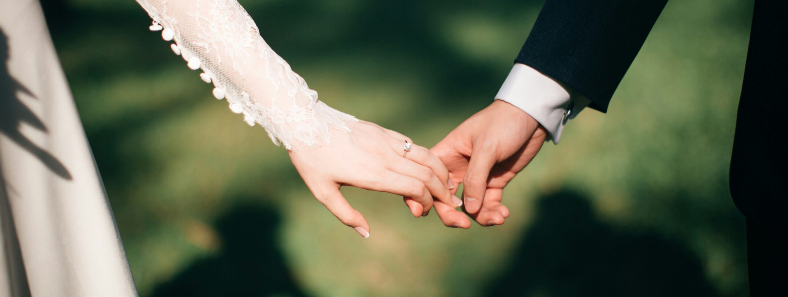 Close-up of a couple holding hands. The woman's hand has a lace sleeve, while the man's hand is in a dark suit. The background is a blurred green outdoor scene.