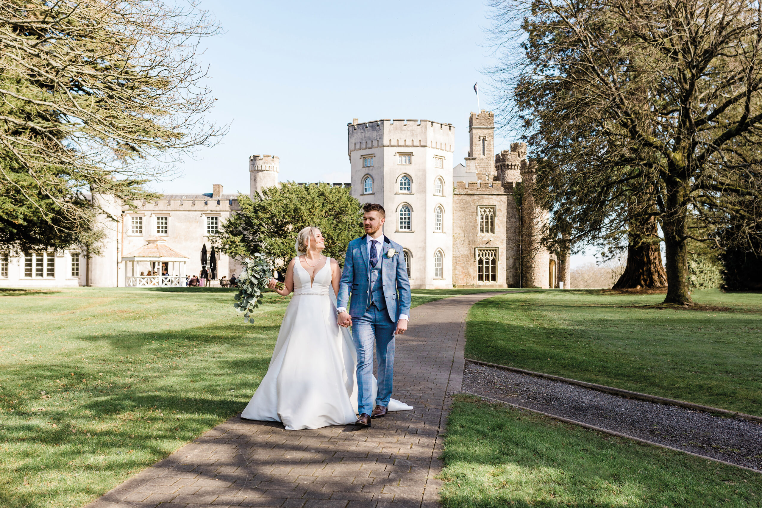 A couple in wedding attire walks hand in hand along a path in front of a large castle.