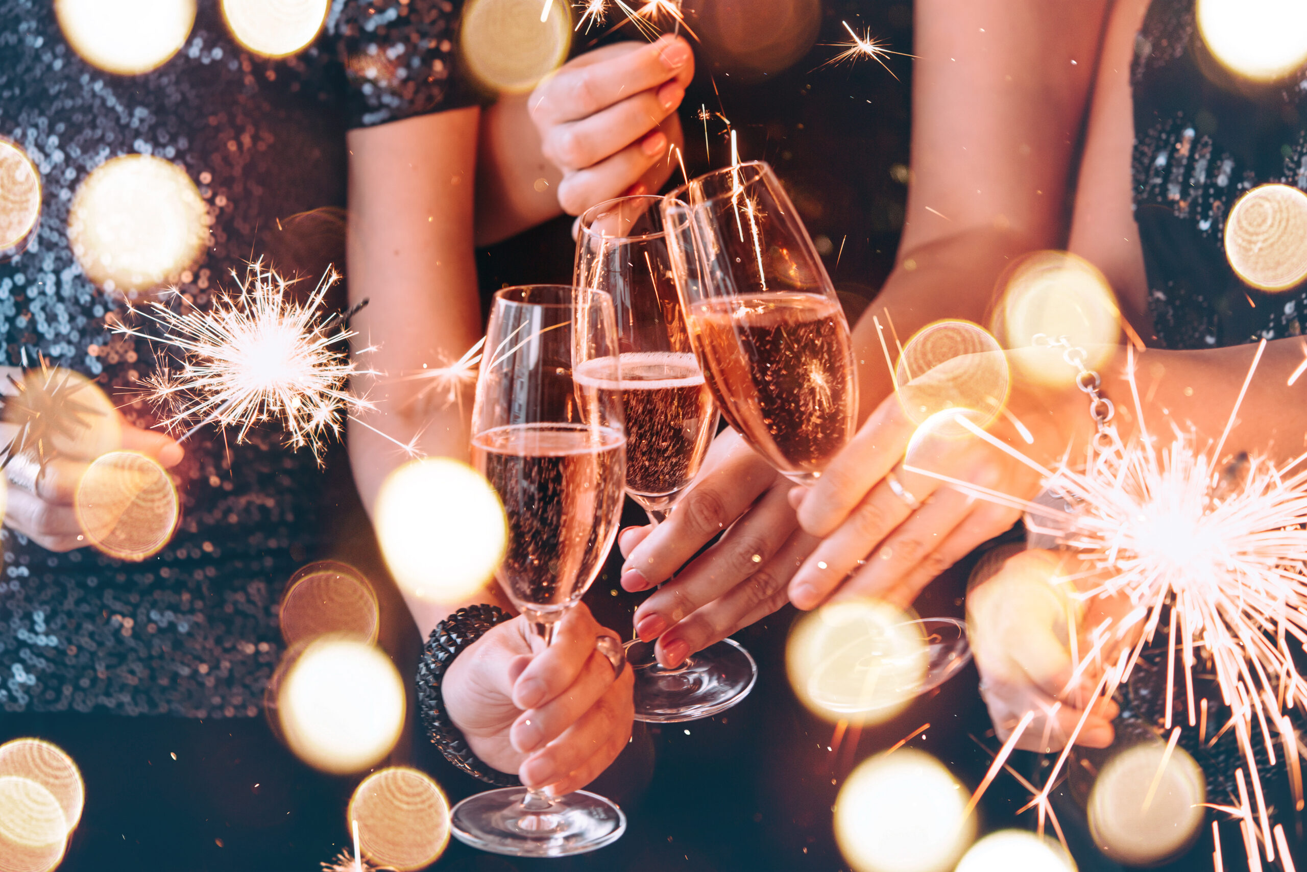 Four people clink glasses of sparkling wine, surrounded by sparklers and festive lights.