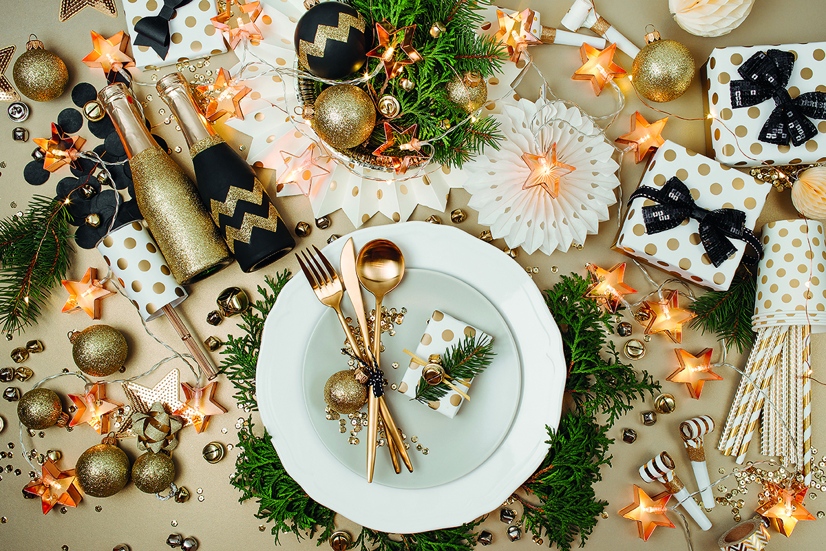 Festive table setting with a white plate, gold cutlery, decorative ornaments, pine branches, wrapped gifts, and string lights in star shapes.