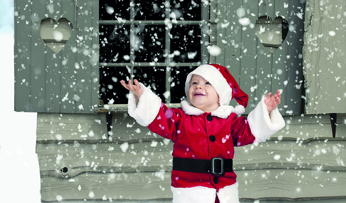 A child in a Santa outfit joyfully catches falling snowflakes outside a wooden house with heart-shaped window shutters.