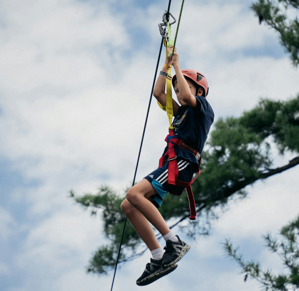 A person wearing a helmet and harness rides a zipline against a background of trees and sky.