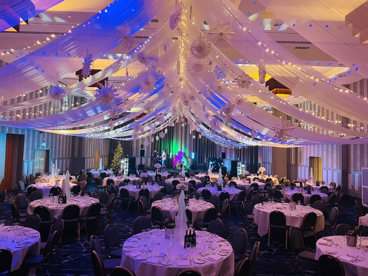 A large banquet hall decorated with draped white fabric and string lights, set for an event with round tables and chairs. A Christmas tree and stage with lights are visible in the background.