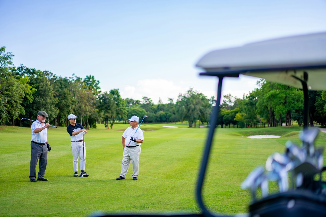 Three people stand on a green golf course, holding clubs and talking, with a golf cart and trees in the background.