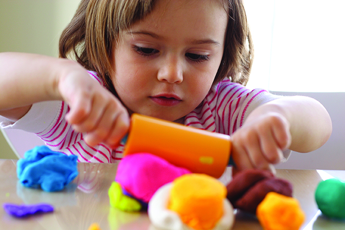 A child intently molds colorful clay with a plastic tool on a table.