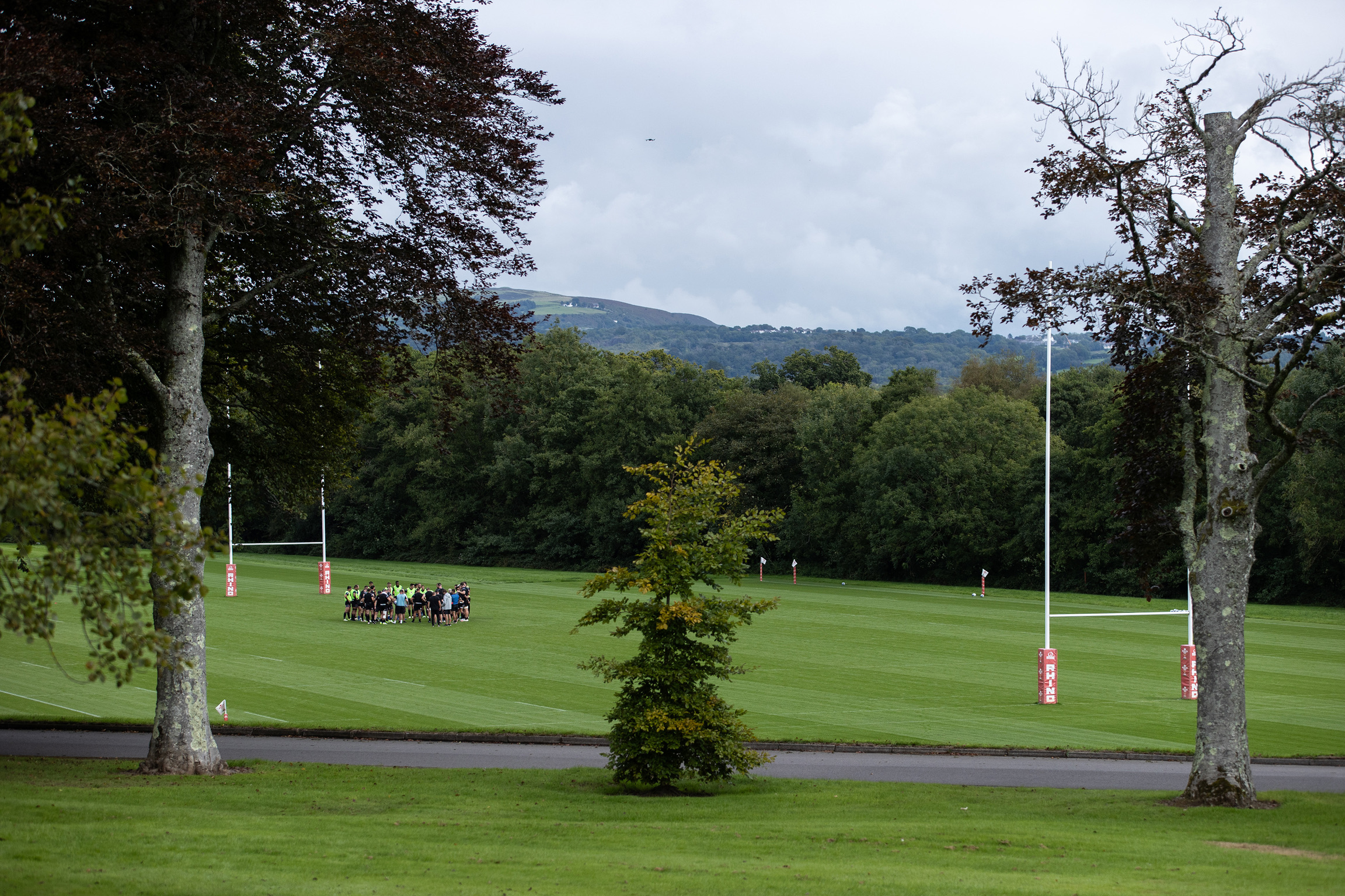 A group of people gather on a rugby field surrounded by trees, with goalposts visible and hills in the background under a cloudy sky.