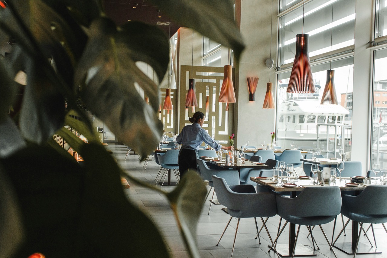 A restaurant with modern decor; a person arranges tables near large windows, with blue chairs and wooden light fixtures.