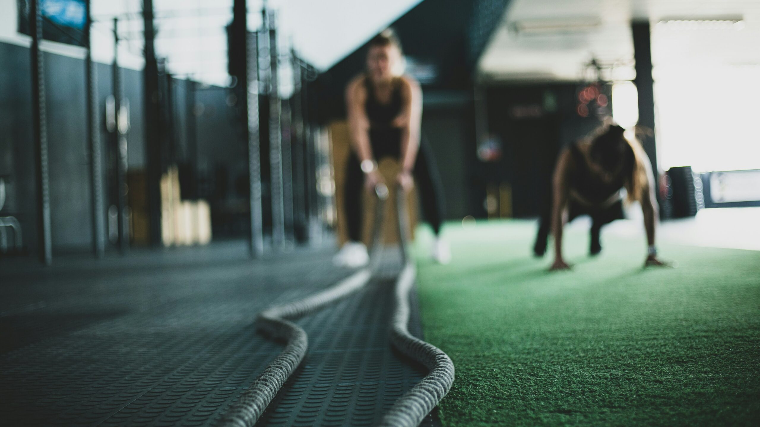 Two people working out indoors. One uses battle ropes, the other appears to be in a push-up position on a green and black floor. The focus is on the ropes in the foreground.