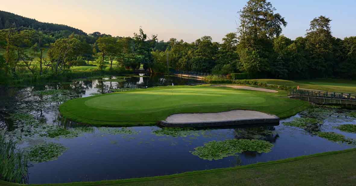 A golf course with a putting green surrounded by water and trees, featuring a sand trap on the right, under a clear sky.