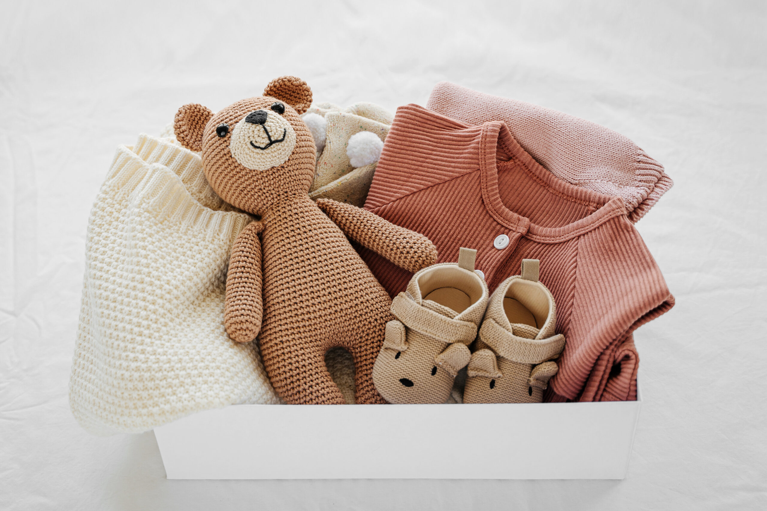 A gift box containing baby clothes, a knitted teddy bear, cream and pink-colored outfits, and a pair of baby shoes on a white background.