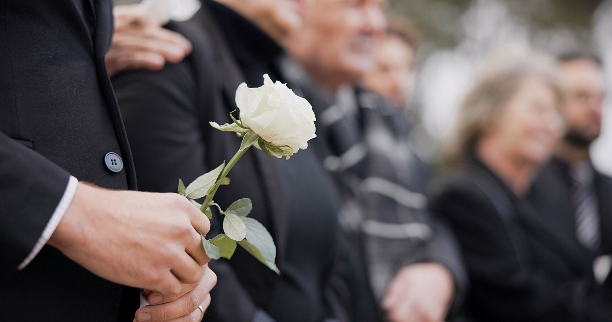 A person in a black suit holds a white rose while standing with others in dark clothing.