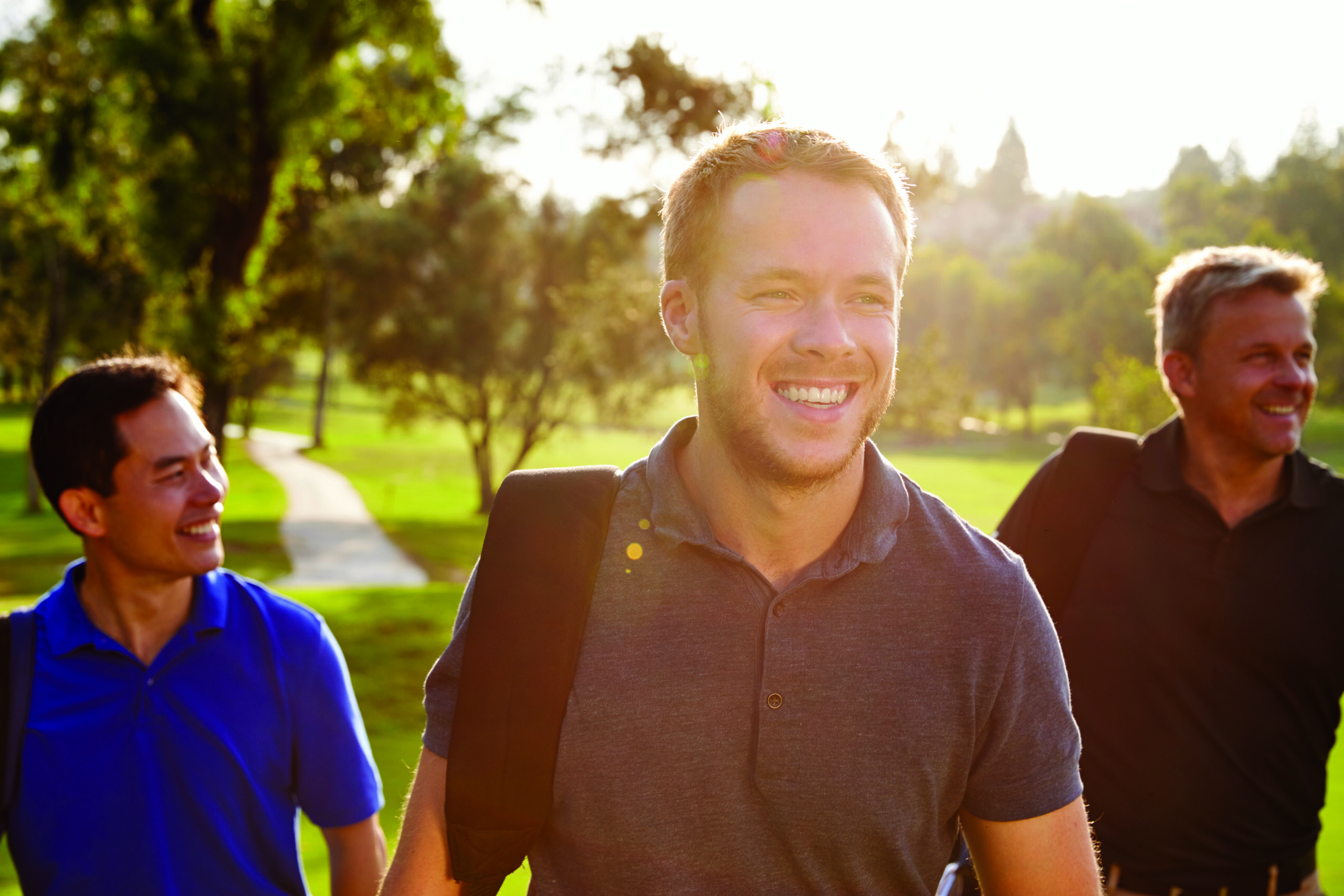 Three men smiling and walking on a sunlit golf course, carrying golf bags over their shoulders.