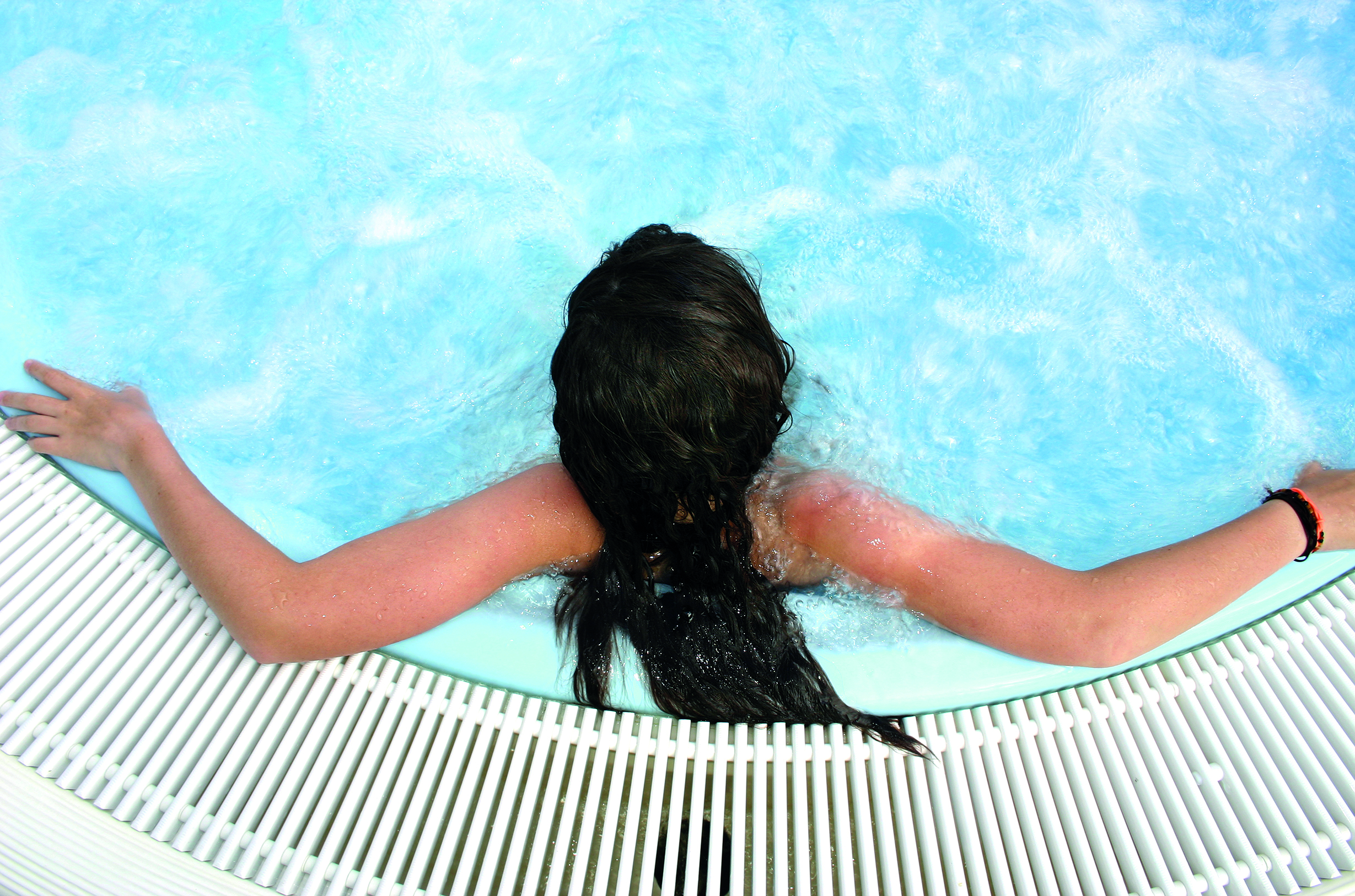 Person with long dark hair relaxing in a pool, arms resting on the pool edge, surrounded by white slats.