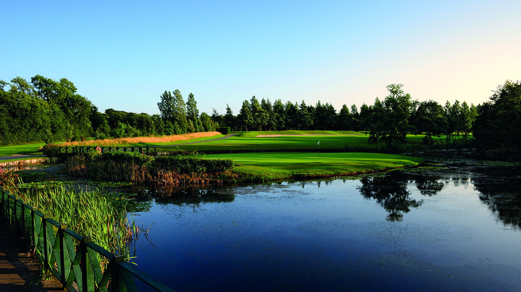 A scenic golf course with lush green fairways, a still pond, and trees in the background under a clear blue sky.