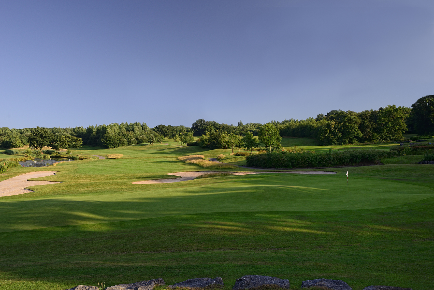 A green golf course with sand bunkers and neatly trimmed grass on a clear day. Trees line the course in the background.