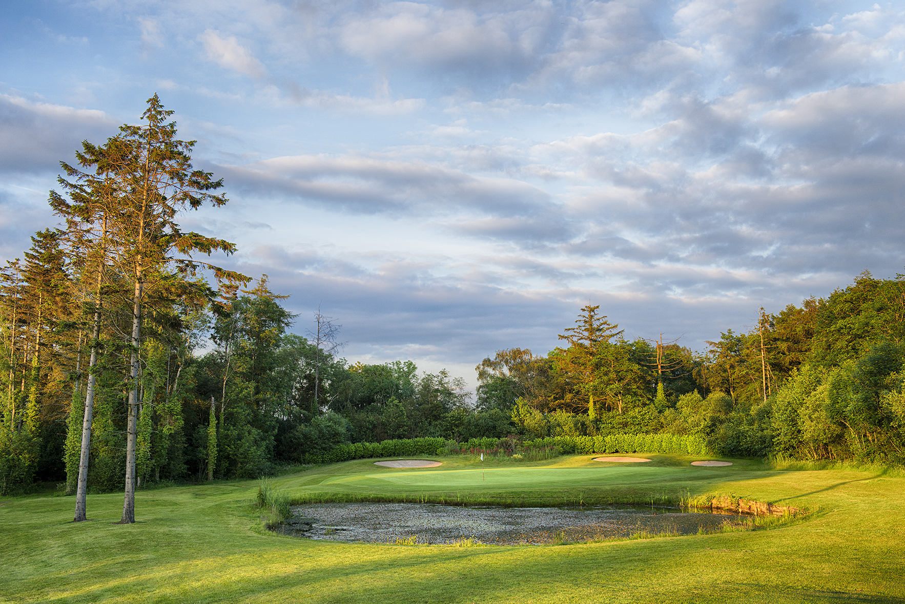 Golf course landscape with a small pond, sand traps, and surrounding trees under a partly cloudy sky.