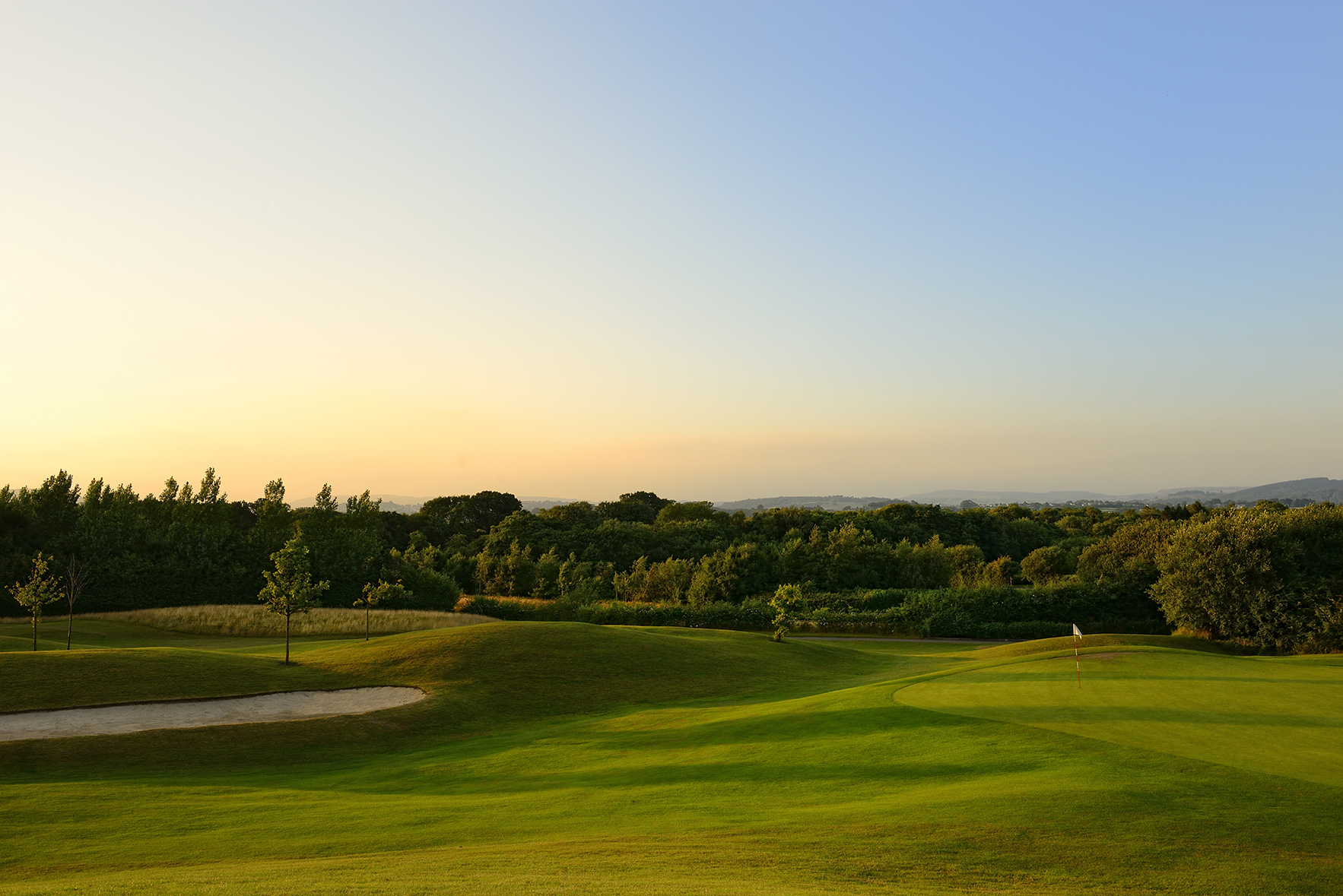 A golf course with a sand trap and flagstick under a clear sky at sunrise or sunset. Lush greenery and trees surround the area.