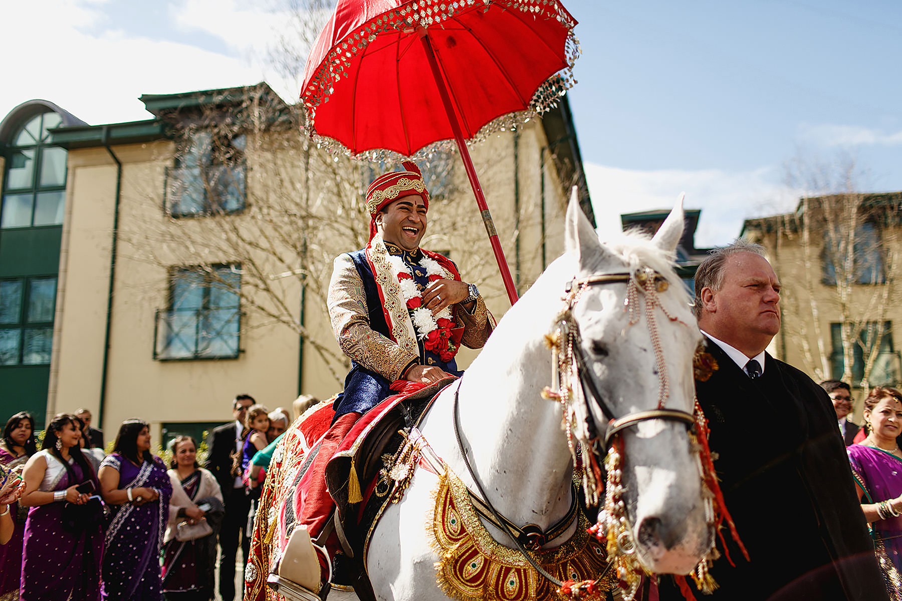 A groom in traditional attire rides a white horse under a red umbrella, leading a procession with guests.