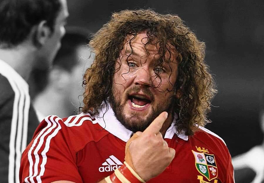 A rugby player with long curly hair and a beard is pointing and speaking, wearing a red jersey with team logos.