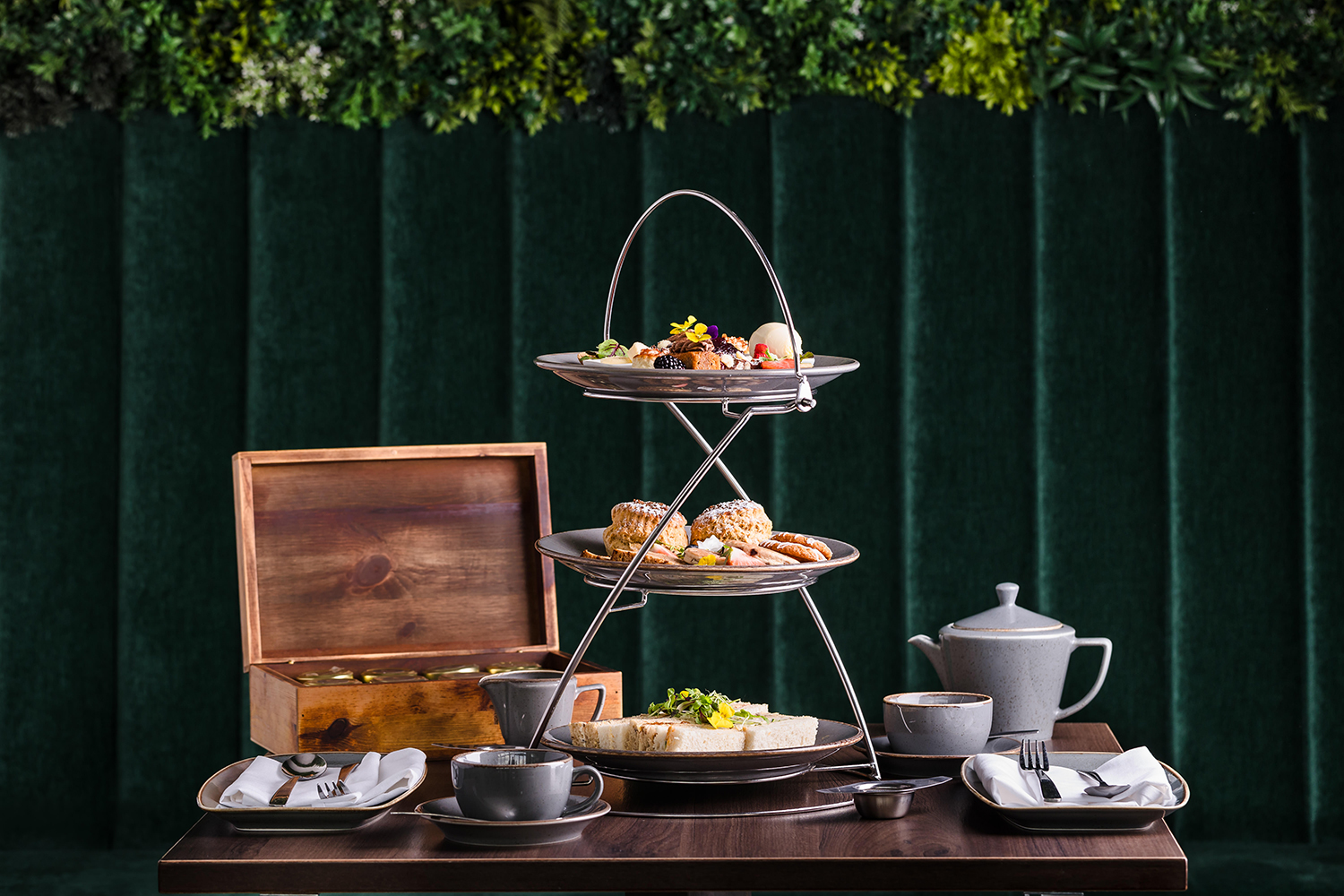 Two-tiered tea stand with assorted pastries and sandwiches, teapot, cups, and wooden box on a table with green backdrop.