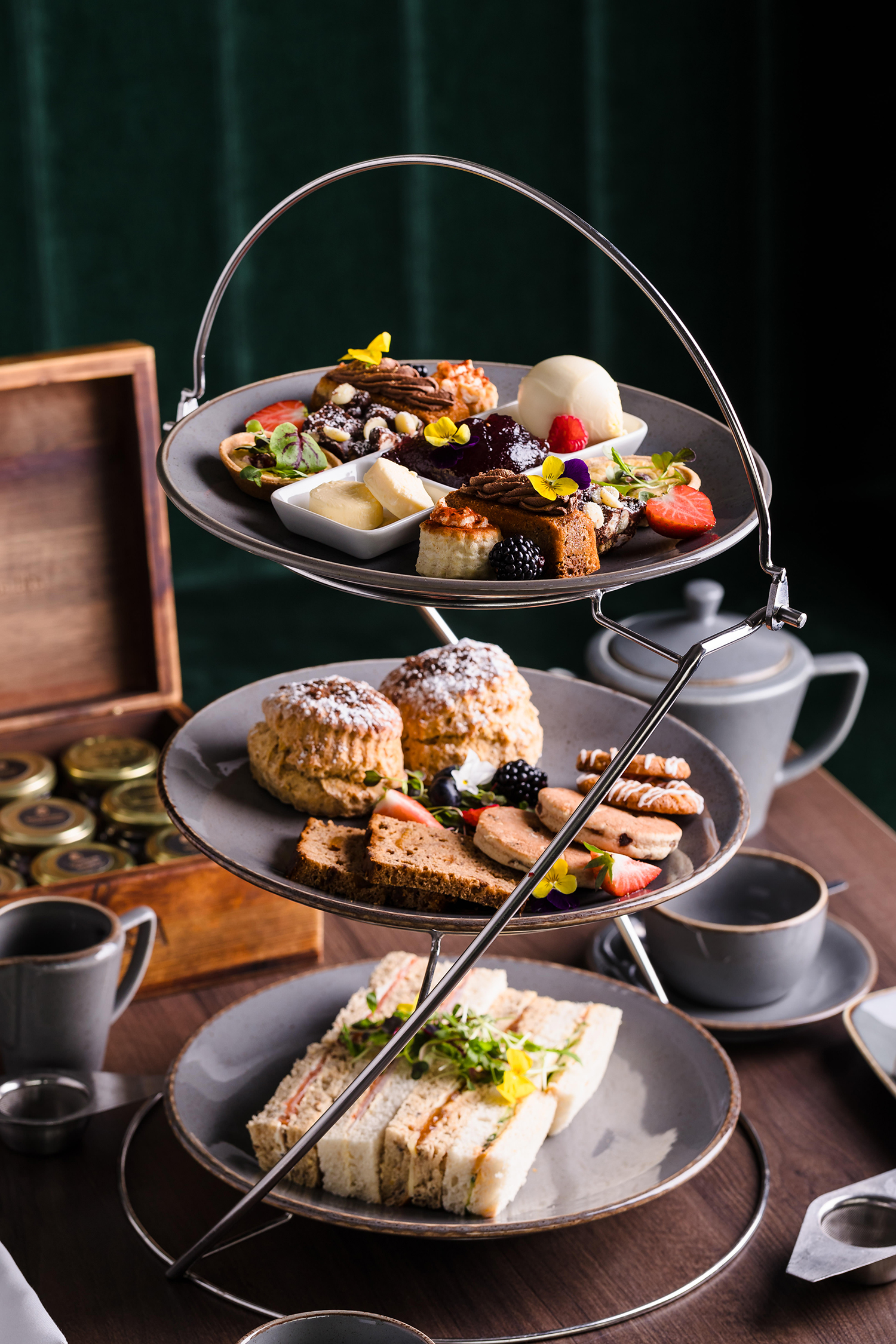 Three-tier stand with assorted pastries, scones, and sandwiches on a table with tea cups and a wooden box.