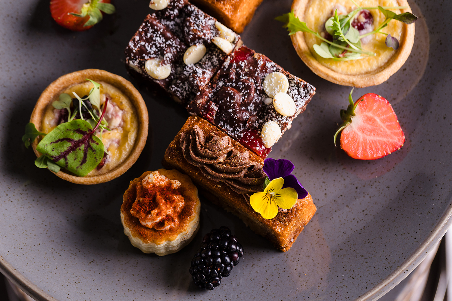 A plate of assorted desserts, including tarts, a chocolate slice with almonds, and fresh berries, garnished with edible flowers.