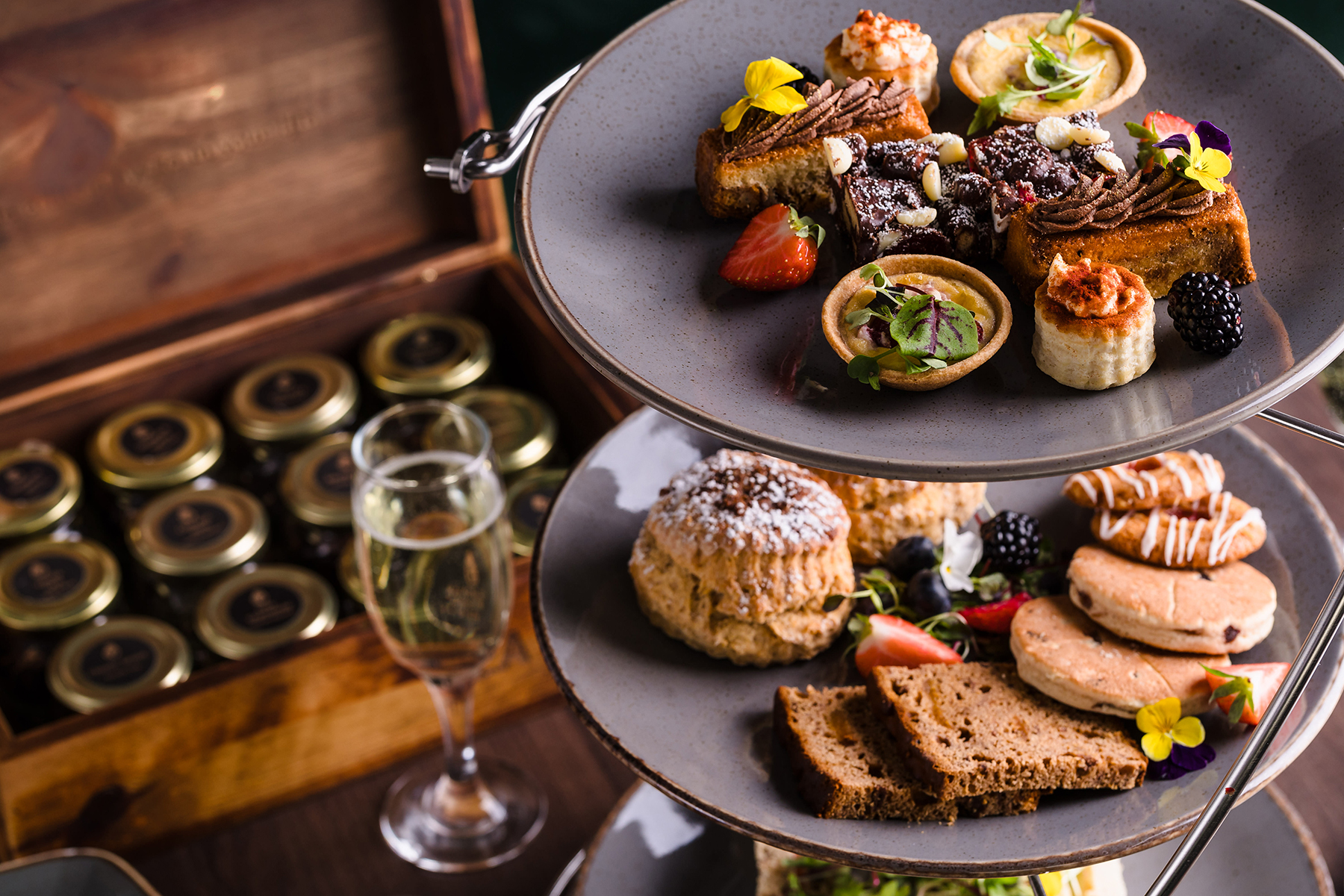 Tiered platter with assorted pastries, fruits, and scones. A wooden box with jars and a glass of champagne are beside it.